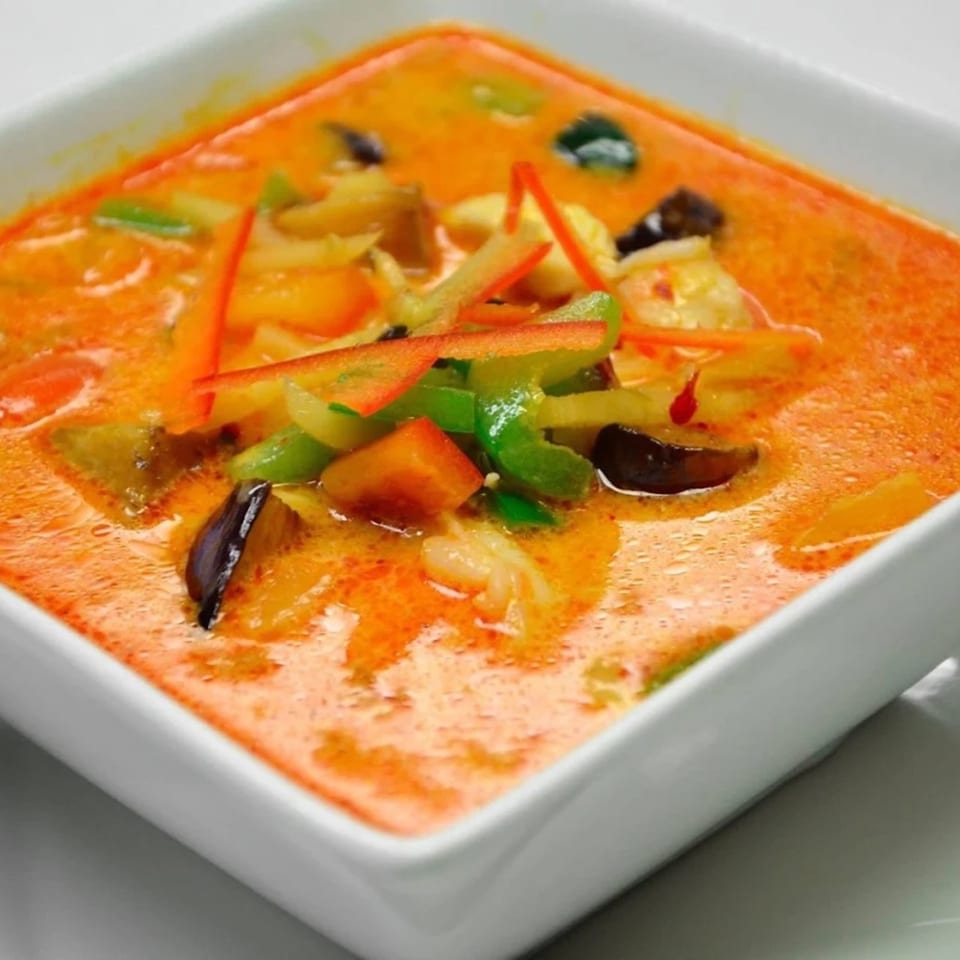 Close-up of Omaha Mercy Thai red curry with vegetables and shrimp in a white square bowl.