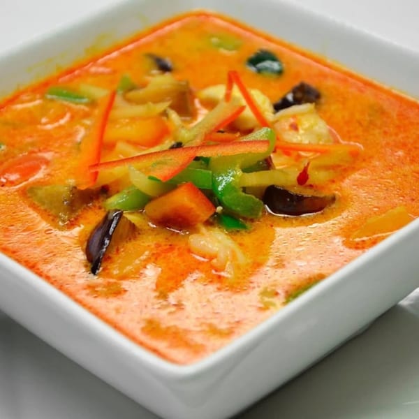 Close-up of a square white bowl filled with vibrant orange Thai curry, vegetables, and shrimp.