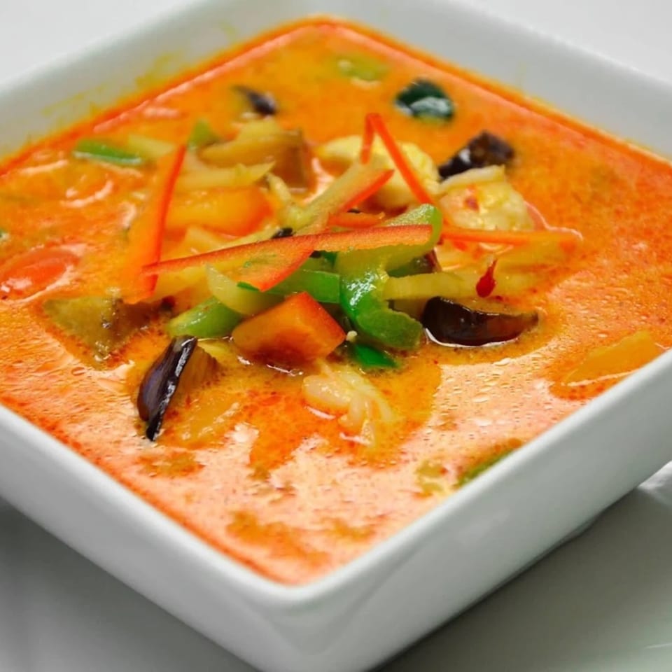 Close-up of a square white bowl filled with vibrant orange Thai curry, vegetables, and shrimp.