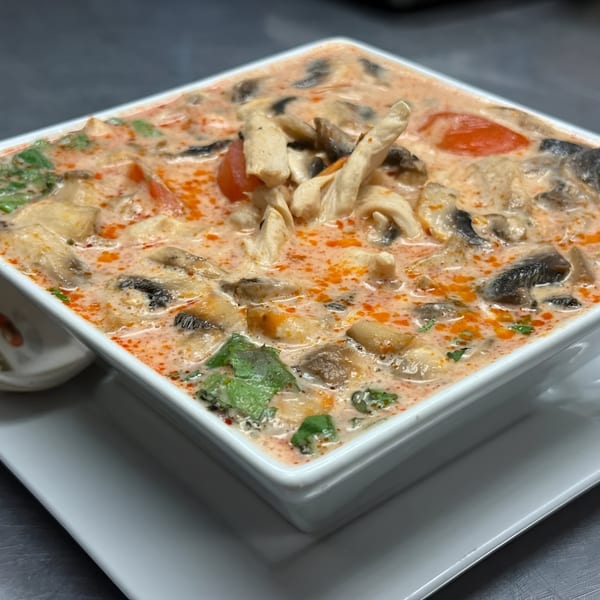 A square white bowl of creamy Tom Kha Gai soup with chicken, mushrooms, and tomatoes.