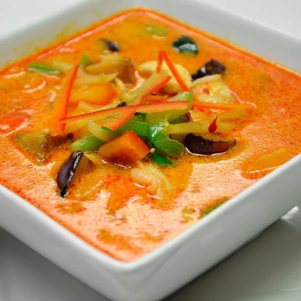 Close-up of a square white bowl filled with vibrant orange Thai curry, garnished with colorful vegetables.