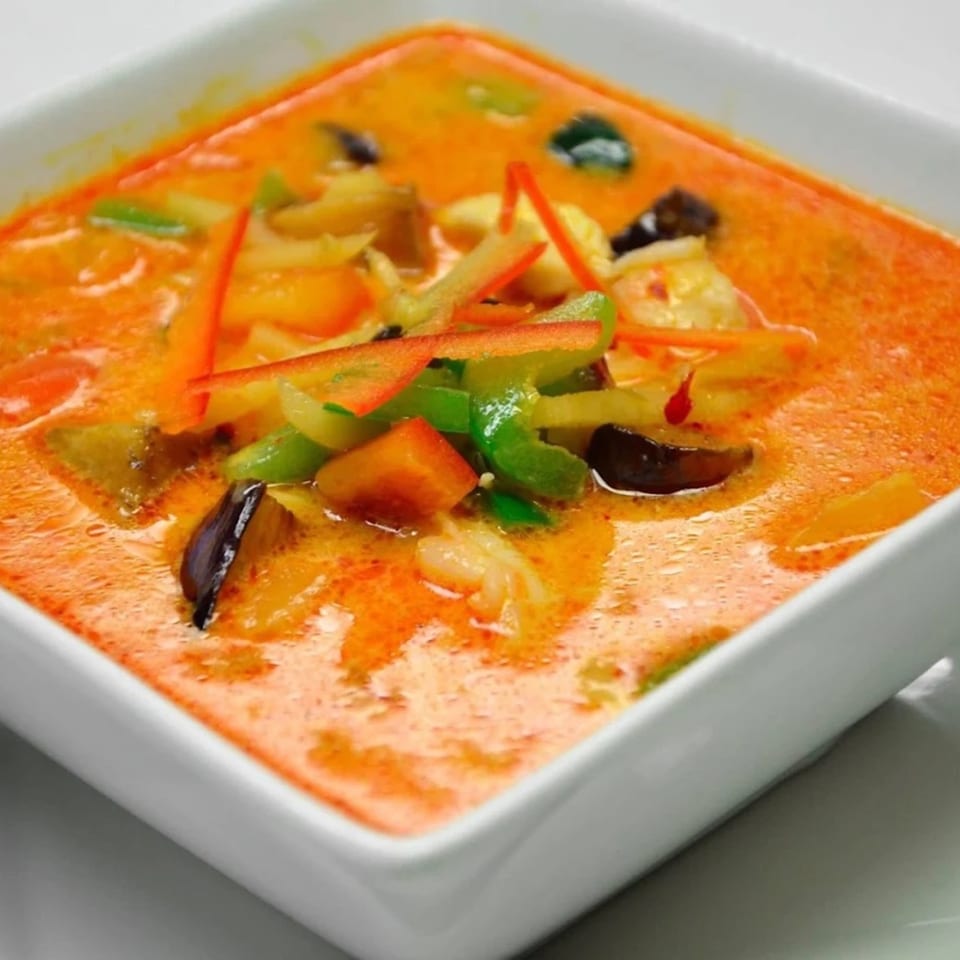 Close-up of a square white bowl filled with vibrant orange Thai curry, garnished with colorful vegetables.
